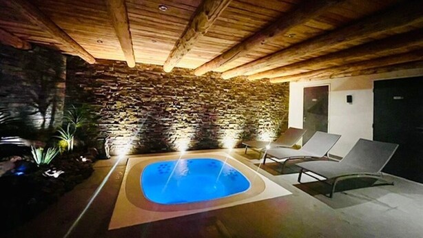 Área interna de piscina com paredes de pedra, teto de madeira, espreguiçadeiras e uma pequena mesa.