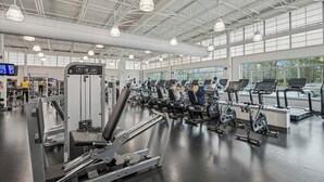 Un gimnasio con diferentes máquinas de ejercicio, amplias ventanas y un techo alto con iluminación industrial.