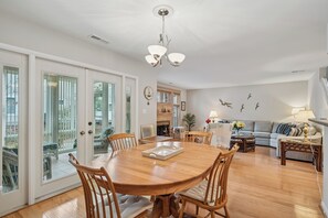 A spacious living room with a wooden dining table, comfortable seating, a fireplace, and a view through large glass doors to an outdoor patio. - Three BR Townhome in Sea Colony (Bethany Beach)