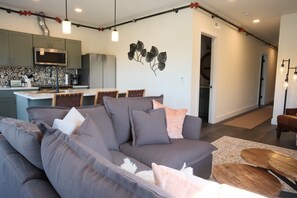 A modern living room with a grey sofa, a dining area with a white table, and a kitchen with grey cabinets and a microwave. - Brand New 2BR River Condo w/ Large Terrace (Indian River)