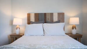 Un lit avec une couette et des oreillers blancs, deux tables de chevet avec des lampes et une tête de lit en bois.
