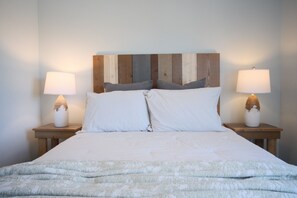 Un lit avec une couette et des oreillers blancs, deux tables de chevet avec des lampes et une tête de lit en bois.