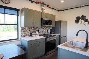 A compact kitchen with a microwave, stove, and sink, a window with a view, and a decorative wall piece. - Brand New 2BR River Condo w/ Large Terrace (Indian River)
