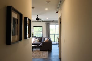 Un couloir mène à un salon équipé d’un canapé, d’un ventilateur de plafond et d’une grande fenêtre.