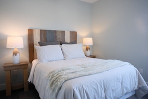 Un lit bien fait, avec une literie blanche et un couvre-lit de couleur claire, flanqué de deux tables de chevet avec des lampes et d’une tête de lit en bois.