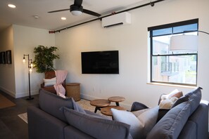 A modern living room with a sofa, a small table, a floor lamp, a plant, and a TV mounted on the wall. - Brand New 2BR River Condo w/ Large Terrace (Indian River)