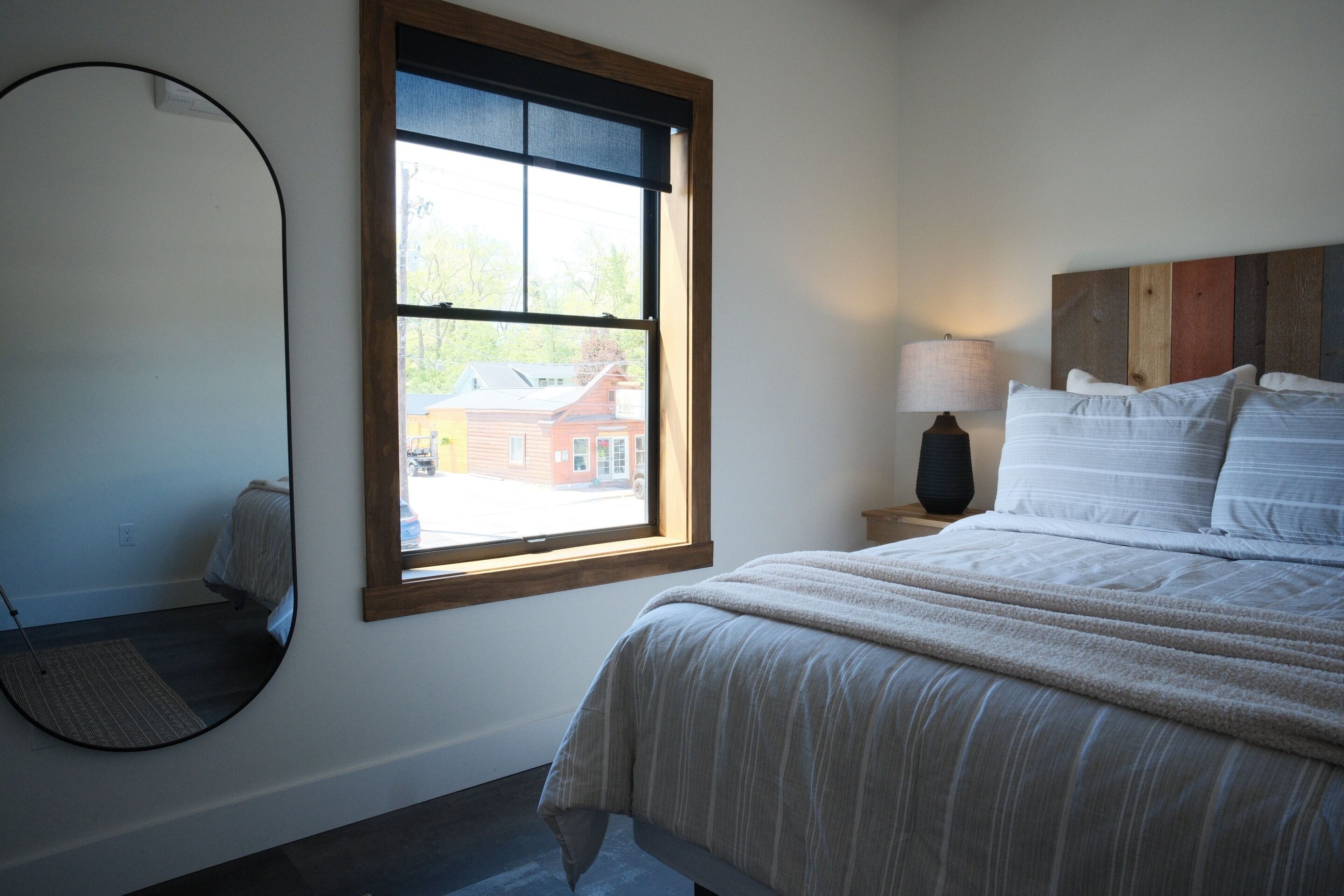 A bedroom with a bed, a round mirror, a window, and a nightstand with a lamp.