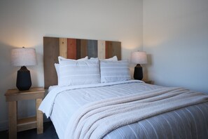 Un lit bien fait, avec du linge de lit blanc et une couverture beige moelleuse, flanqué de deux tables de chevet avec des lampes, le tout adossé à un mur orné d’une tête de lit en panneaux de bois.
