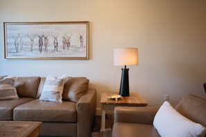 A living room with a brown leather sofa, a wooden side table with a lamp, and a framed picture on the wall. - New 2BR River Condo w/ Balcony (Indian River)