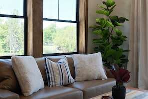 A living room with a sofa, pillows, a plant, and a window with a view of trees.