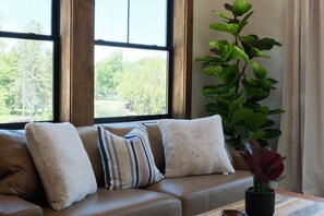 A living room with a sofa, pillows, a plant, and a window with a view of trees. - New 2BR Condo w/Balcony Above Inn Between (Indian River)