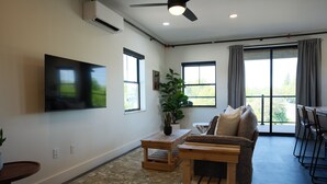 Een woonkamer met een flatscreen televisie, een houten salontafel, een bank, een potplant en een groot raam met gordijnen.