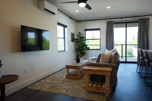 A living room with a flat-screen TV, a wooden coffee table, a sofa, a potted plant, and a large window with curtains.