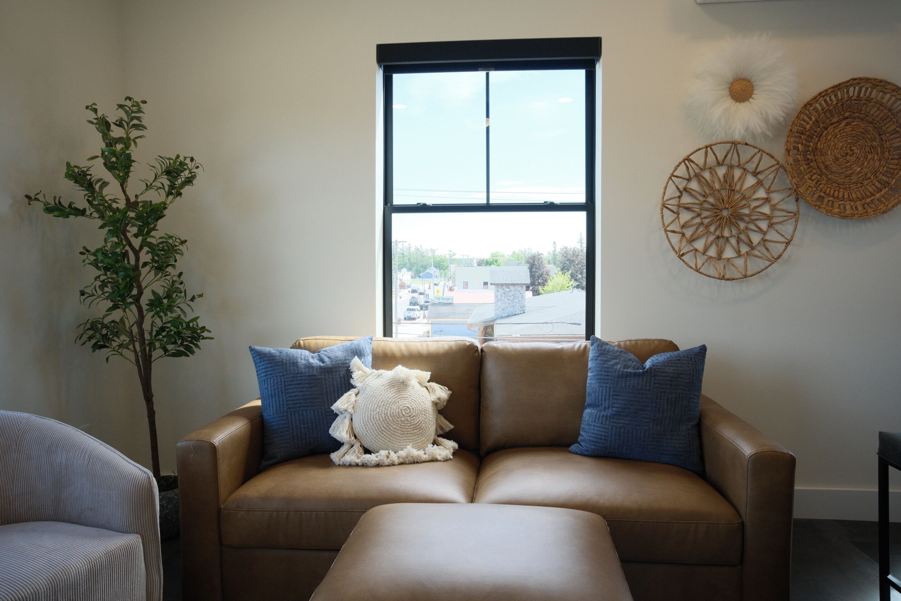 A living room with a brown leather sofa, a potted plant, and a large window with a view of buildings.