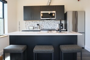 A modern kitchen with dark cabinets, a built-in microwave, a stainless steel refrigerator, and a central island with bar stools. - Brand New 2BR Condo Steps to Downtown (Indian River)