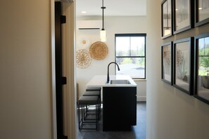 A modern kitchen with a white countertop, black stools, a black sink, and a window with a view of greenery. - Brand New 2BR Condo Steps to Downtown (Indian River)