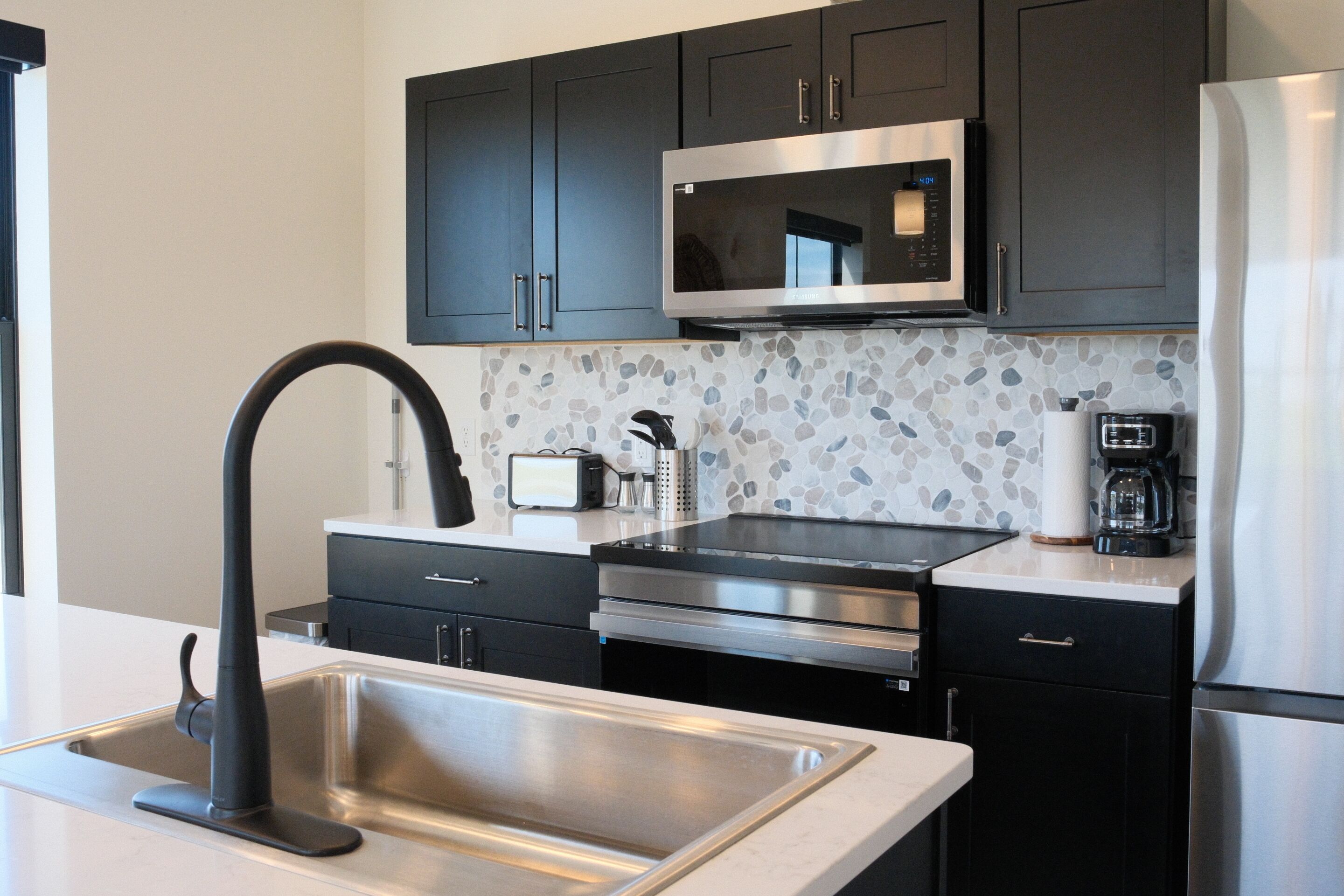 A modern kitchen with dark cabinets, stainless steel appliances, a granite backsplash, and a black faucet.