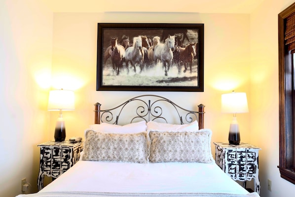 Un lit avec deux tables de chevet, chacune agrémentée d’une lampe et d’une nappe à motifs. Au-dessus du lit, on peut voir un tableau encadré représentant des chevaux.