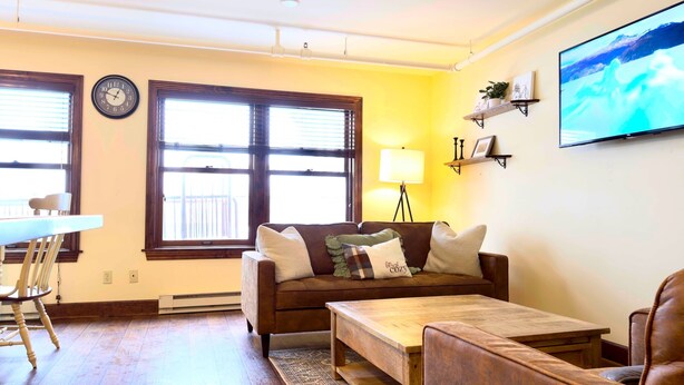 Ein Wohnzimmer mit braunem Sofa, einem Holztisch, einem an der Wand montierten Fernseher und einem Fenster mit Jalousien.