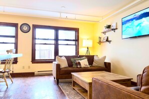 A living room with a brown sofa, a wooden coffee table, a TV mounted on the wall, and a window with blinds. - Astor Street Retreat 3BR Condo (Mackinac Island)