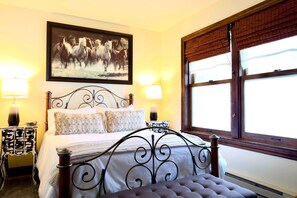 A neatly arranged bedroom with a large bed, two bedside tables with lamps, a framed picture on the wall, and a window with blinds. - Astor Street Retreat 3BR Condo (Mackinac Island)