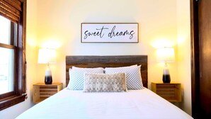 Een net opgemaakt bed met gekleurde kussens, een houten hoofdbord en twee bedlampjes. Een bordje boven het bed staat 'zoete dromen'.
