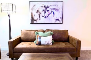 A brown leather sofa with a cushion that reads "let's get cozy" and a framed abstract painting of horses on the wall.