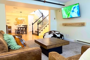 A living room with a wooden coffee table, a brown sofa with green and brown cushions, a large TV mounted on the wall, and a staircase leading to another room.