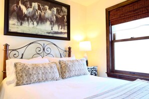 Una cama bien hecha con almohadones estampados, un cuadro enmarcado de caballos encima y una ventana con marco de madera.