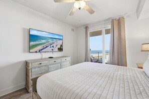 A bedroom with a large bed, a TV on the wall, a ceiling fan, and a view of the beach through the window. - Bourbon Sunsets - WALK to Pier Park, BEACH Access (Panama City Beach)