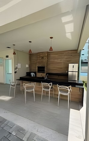 A modern kitchen with a wooden accent wall, stainless steel appliances, and white chairs. - 1501 Apto2 Qtos Vista Mar (Cabedelo)
