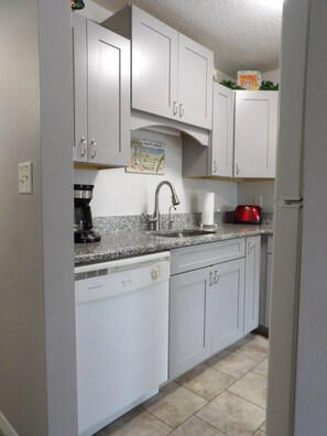 A compact kitchen with white cabinets, a dishwasher, a coffee maker, and a red kettle. - Gulf Shores - Village by the Gulf 2/2 balcony (Gulf Shores)