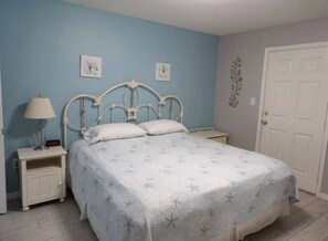 A bedroom with a bed, nightstand, lamp, and a door. - Gulf Shores - Village by the Gulf 2/2 balcony (Gulf Shores)