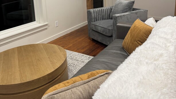 Een moderne woonkamer met een grijze bank, een ronde houten salontafel en een groot raam.