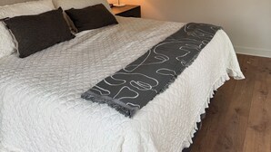 Een slaapkamer met een bed, kussens, een bedlampje, een aan de muur hangend schilderij en een houten vloer.