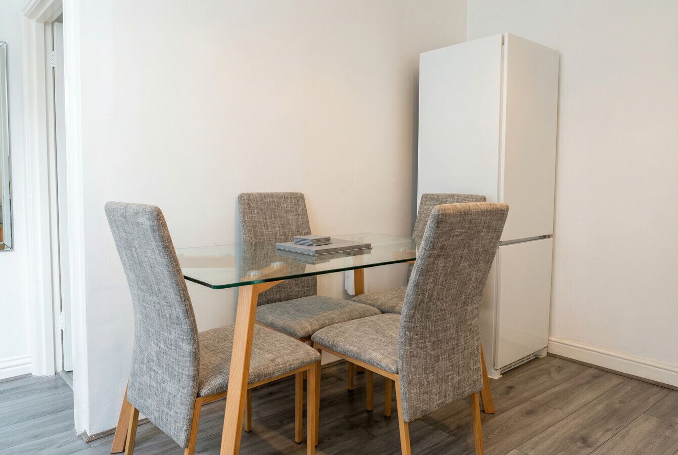 A dining area with a glass table and four grey upholstered chairs, a white refrigerator, and wooden flooring.