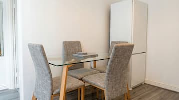 A dining area with a glass table and four grey upholstered chairs, a white refrigerator, and wooden flooring.