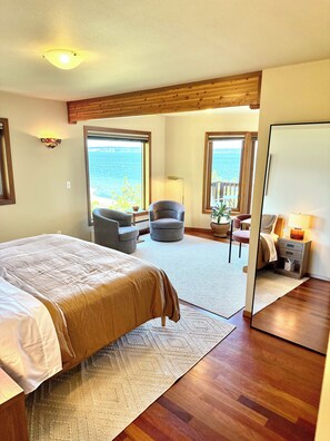 A bedroom with a large bed, a chair, a small table, and a view of the sea through large windows.