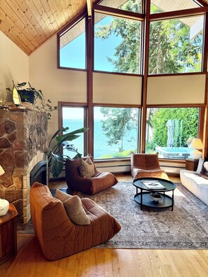 A spacious living room with a stone fireplace, large windows offering a view of the sea, and comfortable seating areas.