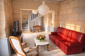 Una sala de estar con un sofá de cuero rojo, una silla de madera, una mesita con un florero, un televisor y una escalera que conduce a otra habitación.
