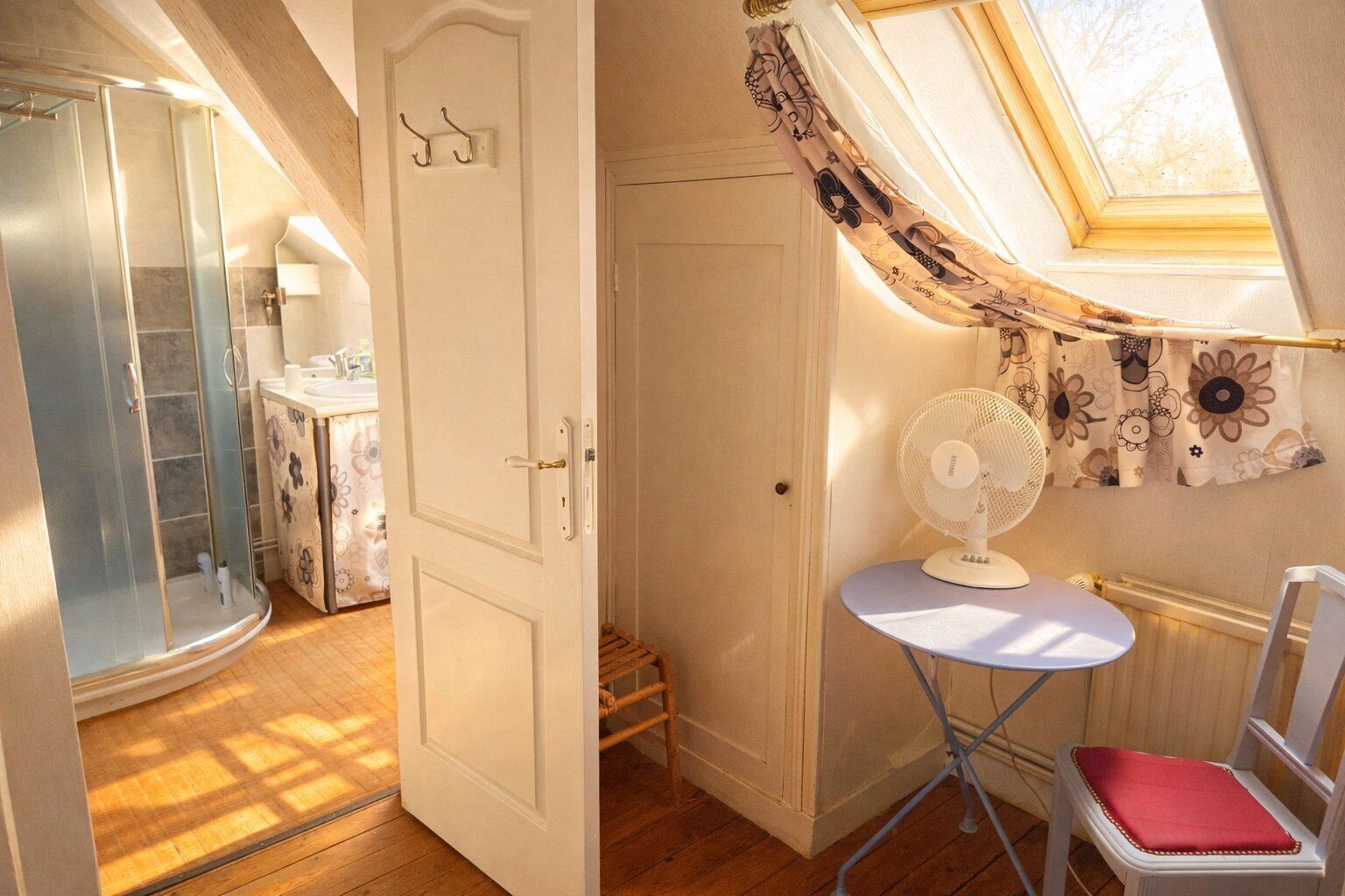 A room with a shower, a small table with a fan, and a chair.