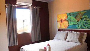A hotel room with a bed, curtains, a painting, and a window. - Apartment in downtown Búzios | 2 bedrooms | Air conditioning (Armação dos Búzios)