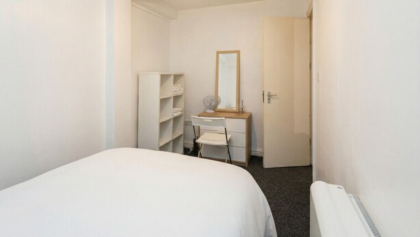 Quarto com cama, cômoda, espelho, ventilador e porta que dá para outro espaço.