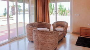 Une pièce meublée de mobilier en osier, dotée d’une porte-fenêtre coulissante et offrant une vue sur des cactus et des palmiers.