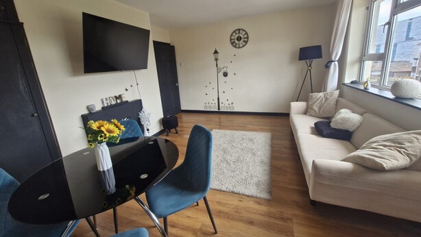 A modern living room with a black dining table, blue chairs, a white sofa, and a TV mounted on the wall. - Apartment in the Heart of Wimbledon  Parking Included! (London)