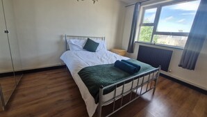 A bedroom with a metal bed frame, white bedding, a green throw blanket, and a window with a view of greenery. - Apartment in the Heart of Wimbledon  Parking Included! (London)