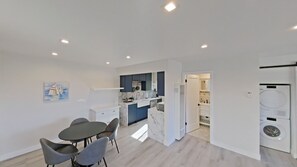 A modern kitchen with dark blue cabinets, white countertops, and a built-in oven. There is a dining area with a table and chairs, and a laundry room with a washing machine. - Beachside Bliss in Carpinteria Blocks to the Beach, Mountain Views (Carpinteria)