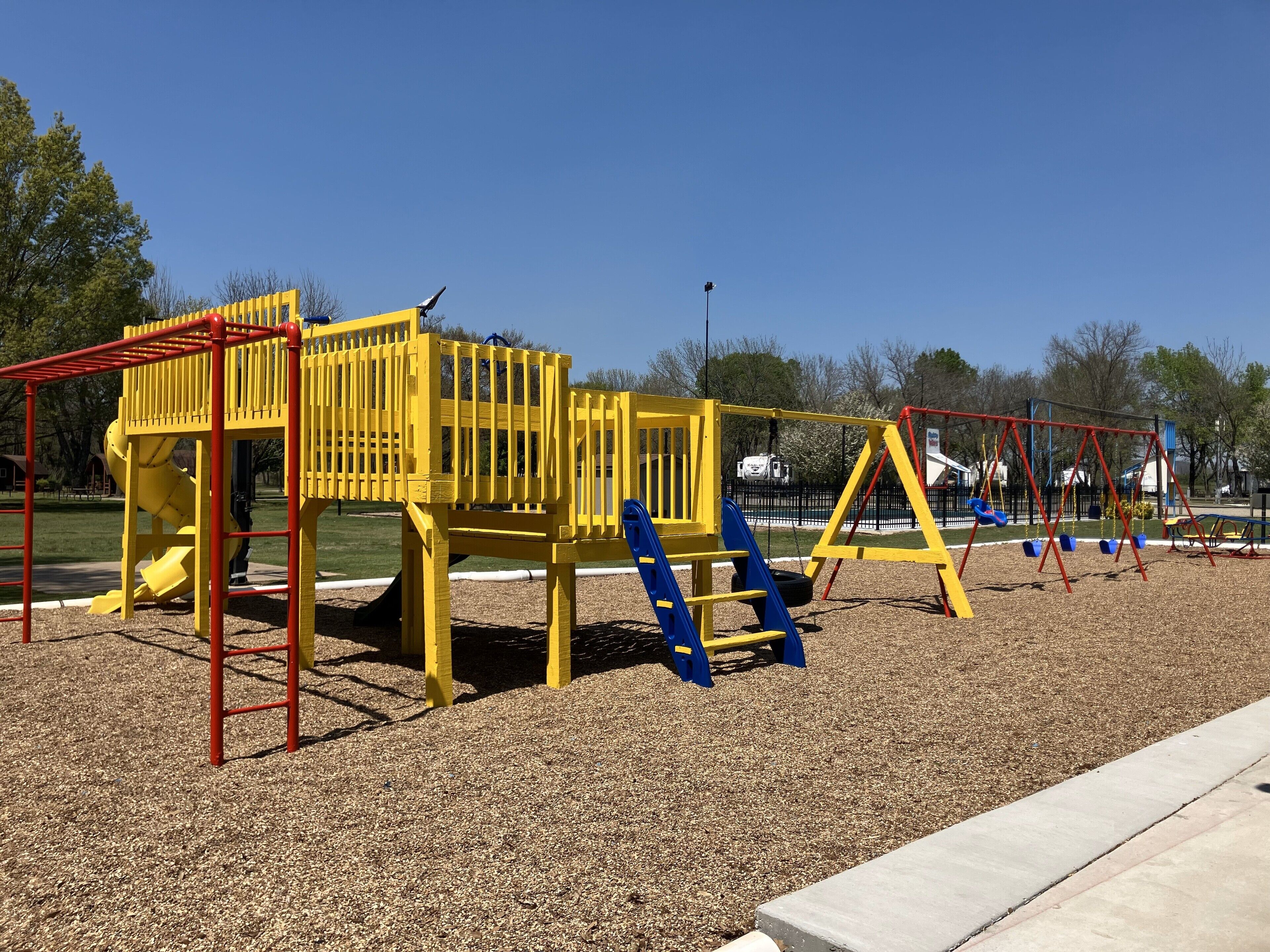Un parque infantil con estructuras de juego en amarillo y rojo, que incluyen un tobogán y columpios, sobre una superficie de grava.