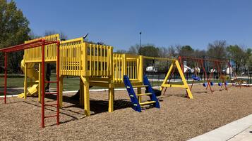 Un parque infantil con estructuras de juego en amarillo y rojo, que incluyen un tobogán y columpios, sobre una superficie de grava.