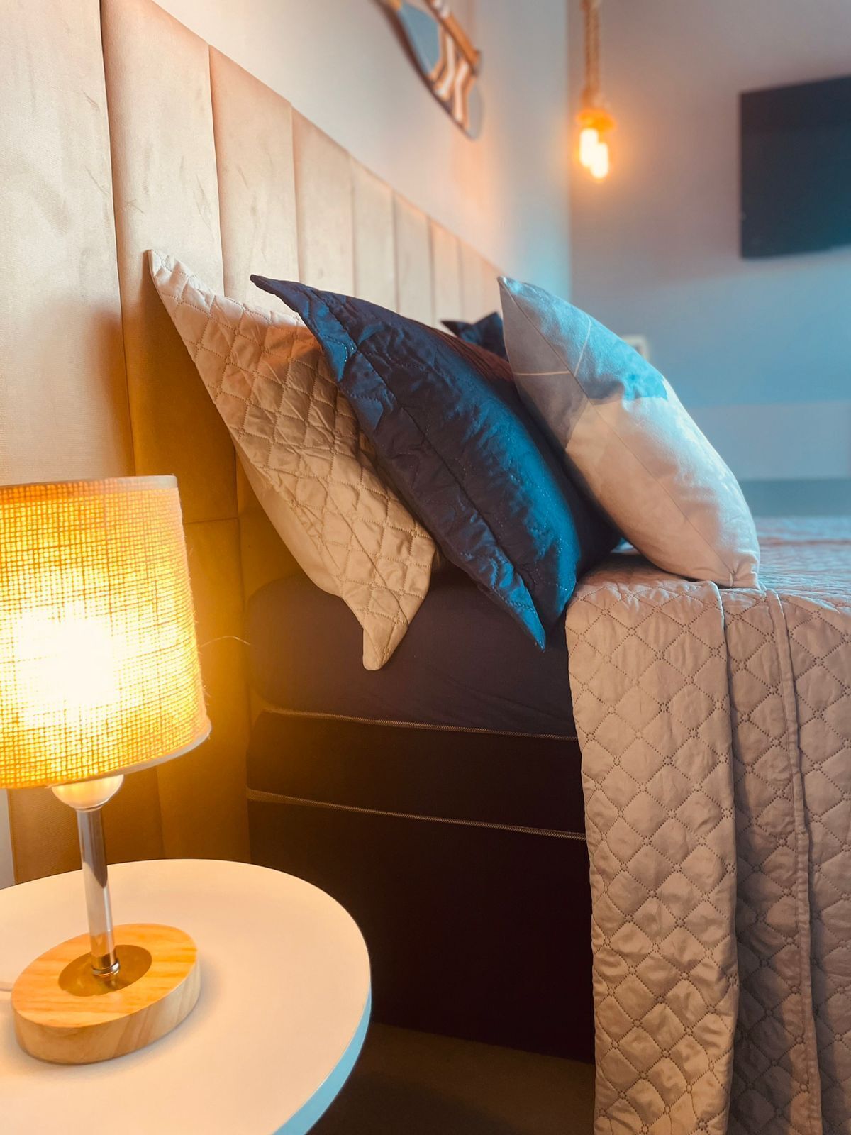 Una cama bien hecha con un cabecero acolchado, una mesita de noche redonda con una lámpara y un televisor fijado a la pared.